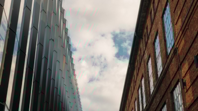 Närbild uppåt mellan en modern glasbyggnad och en äldre tegelfasad under delvis molnig himmel.