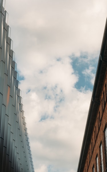 Närbild uppåt mellan en modern glasbyggnad och en äldre tegelfasad under delvis molnig himmel.