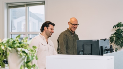 Två personer står framför en Lenovo-skärm på ett kontor med växter och ett fönster med delvis öppna persienner.