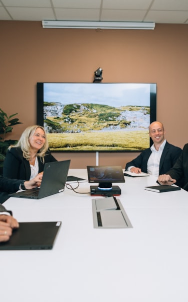 Sex personer i affärskläder sitter runt ett konferensbord i ett modernt kontor och deltar i ett möte. En stor skärm visar ett naturskönt landskap med kullar och grönska.