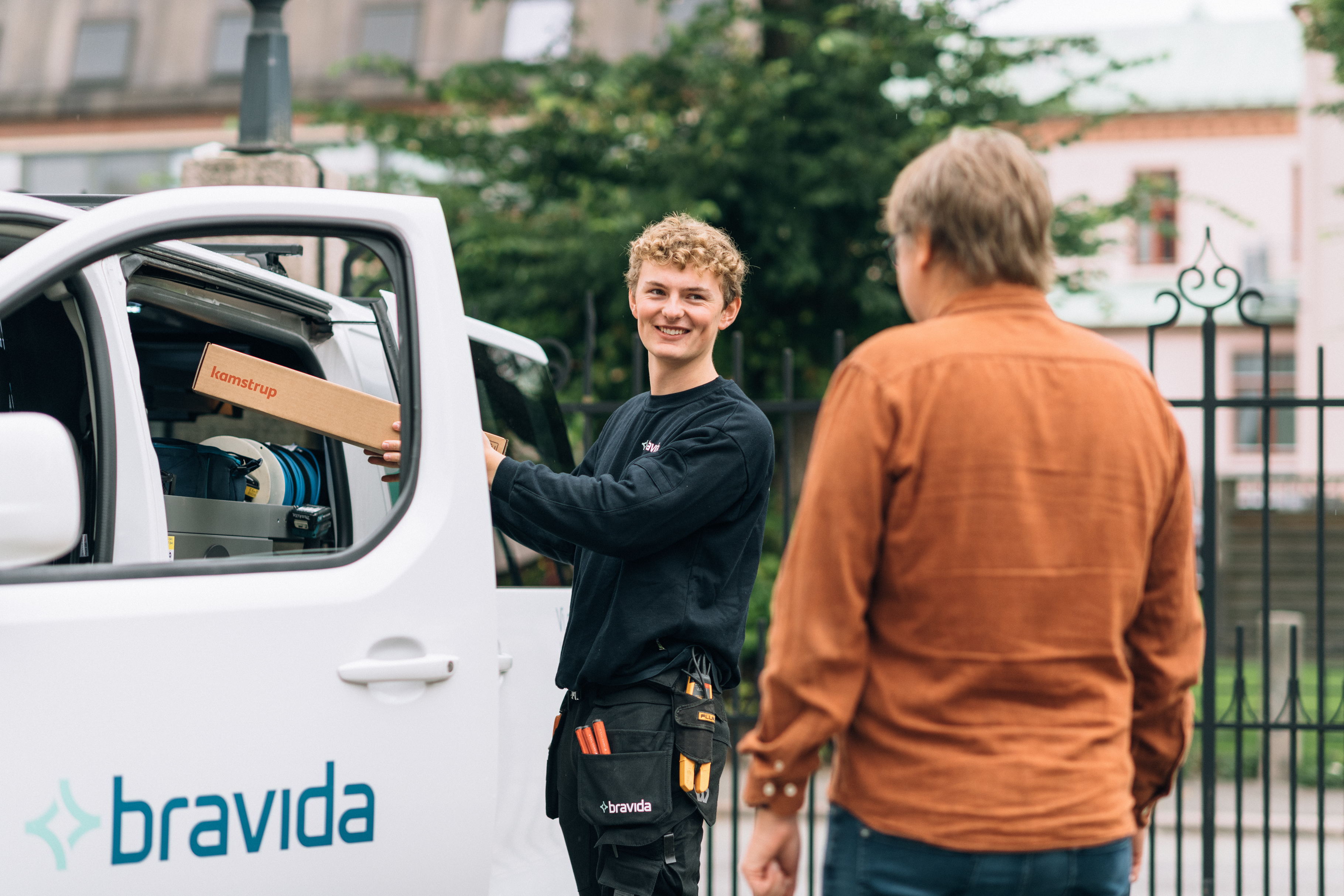 Two people interact near a white van with the Bravida logo. One, wearing a black shirt and tool belt, holds a box labeled "Kamstrup" and appears to hand it to the other, who wears an orange shirt.