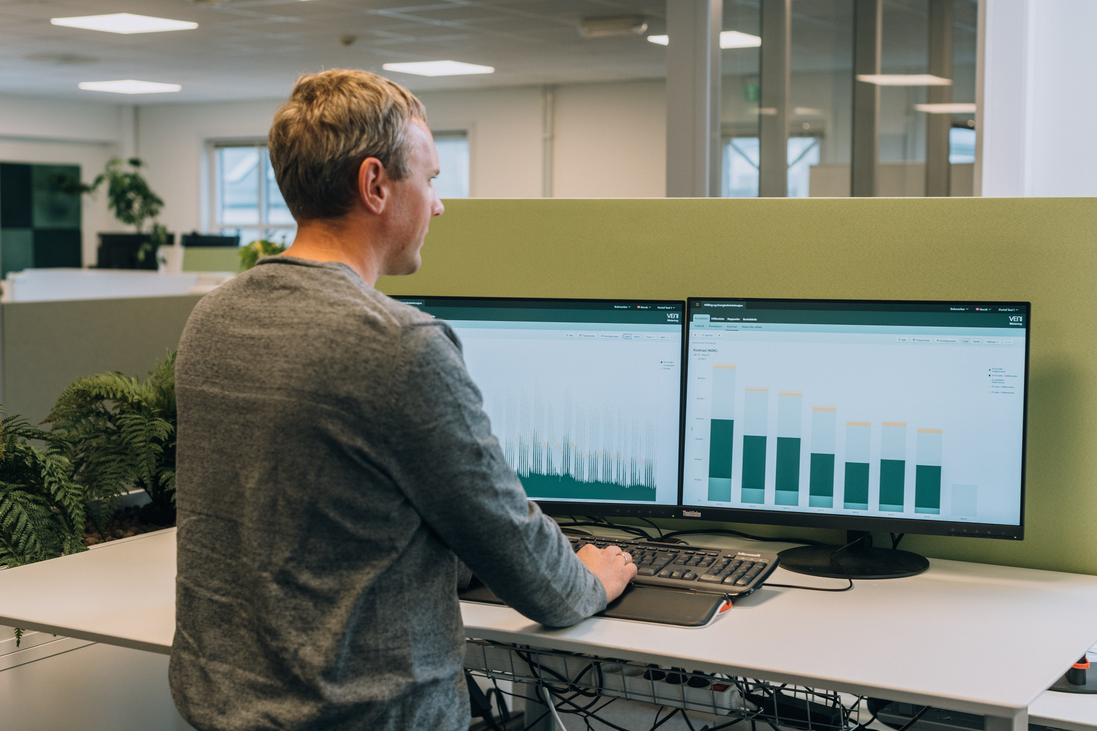 A man stands at a desk in an office, working on two monitors displaying graphs and charts from the VENI Customer Portal.