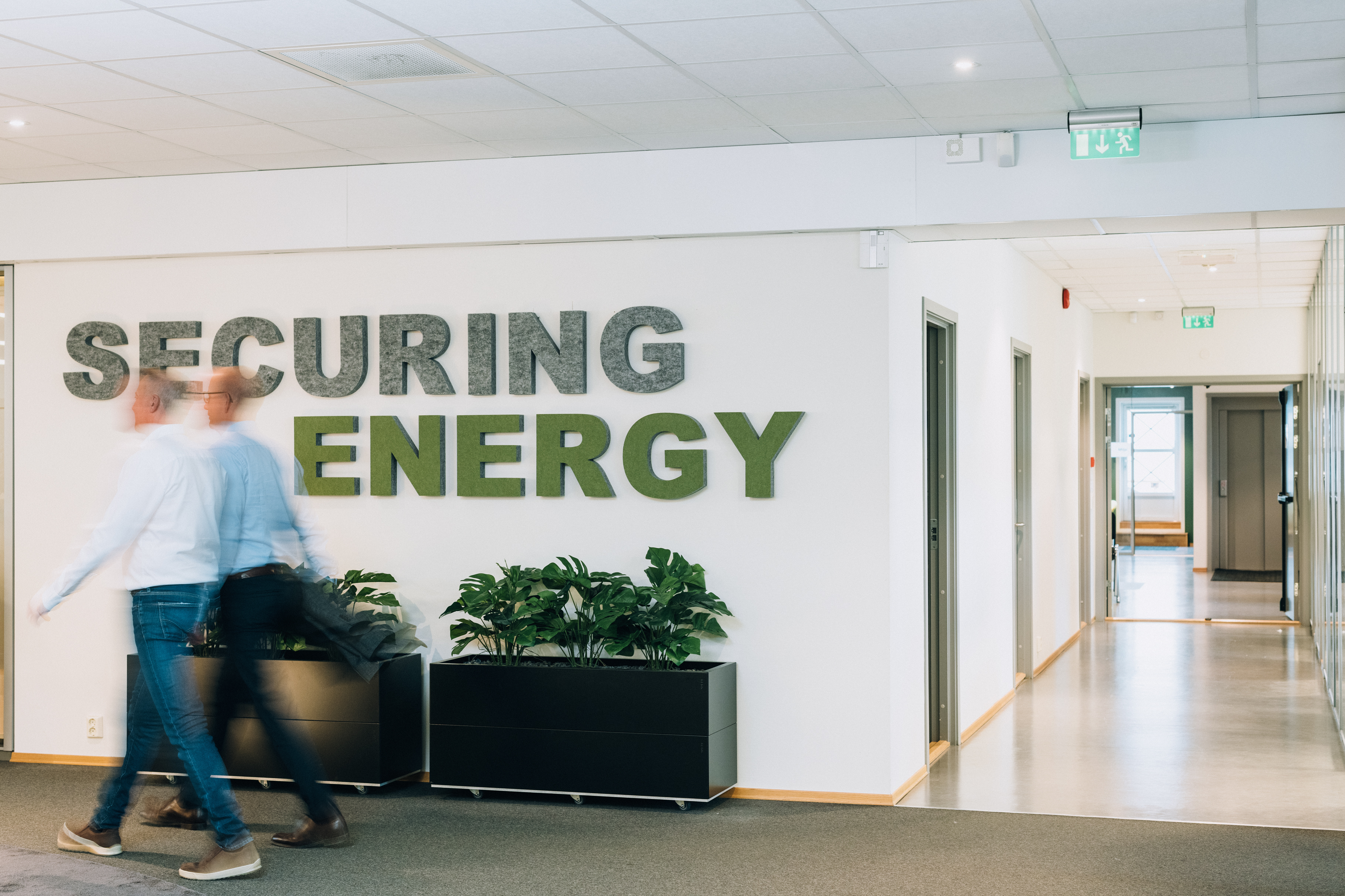 Two people walk through an office hallway with a "SECURING ENERGY" sign on the wall and potted plants below it.