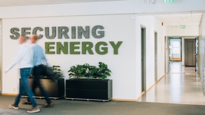 Två personer går genom en kontorskorridor med skylten "SECURING ENERGY" på väggen och krukväxter under.