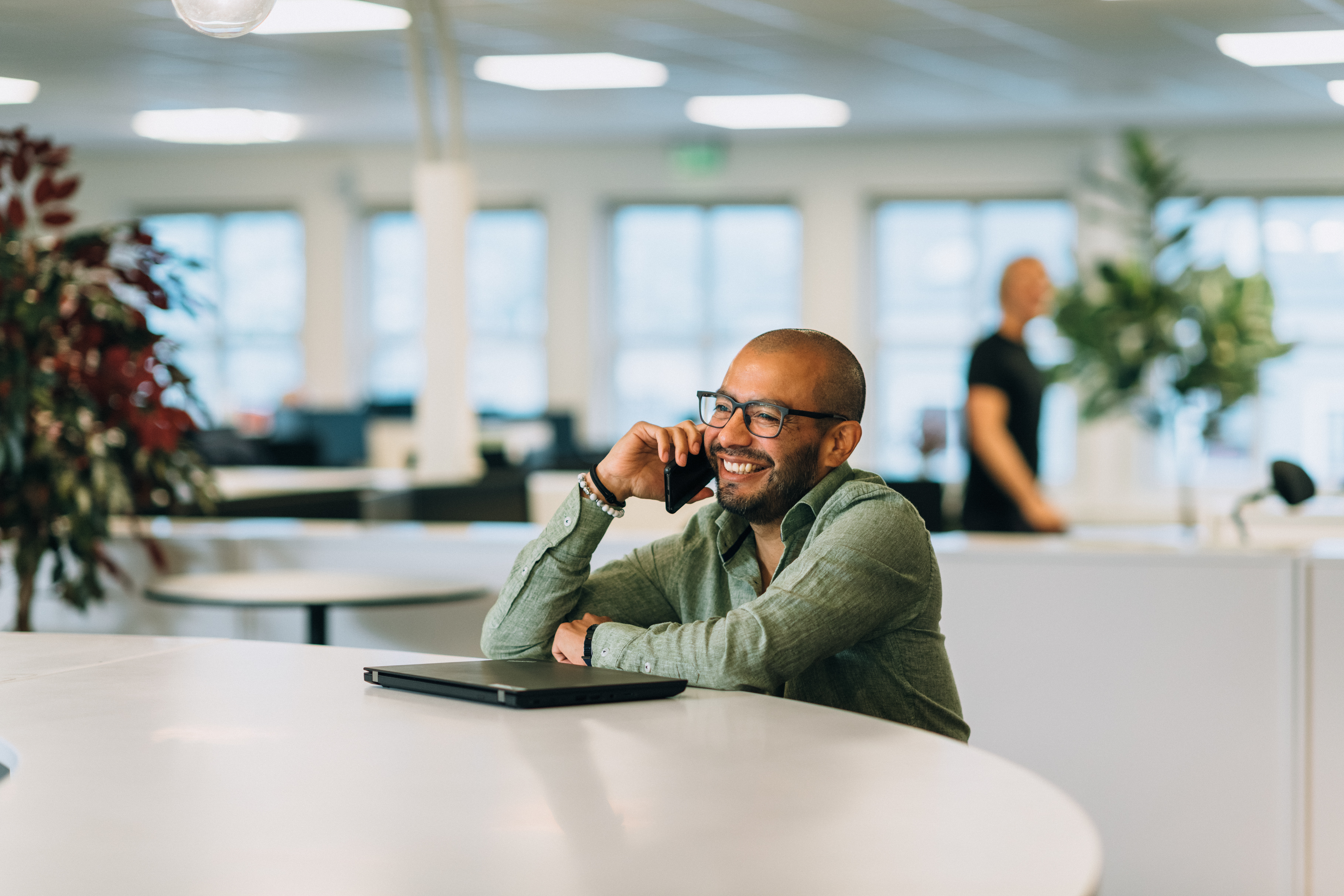 A man sits at a round table in a bright office, holding a phone to his ear. A closed laptop is on the table. A blurred person is walking by in the background.