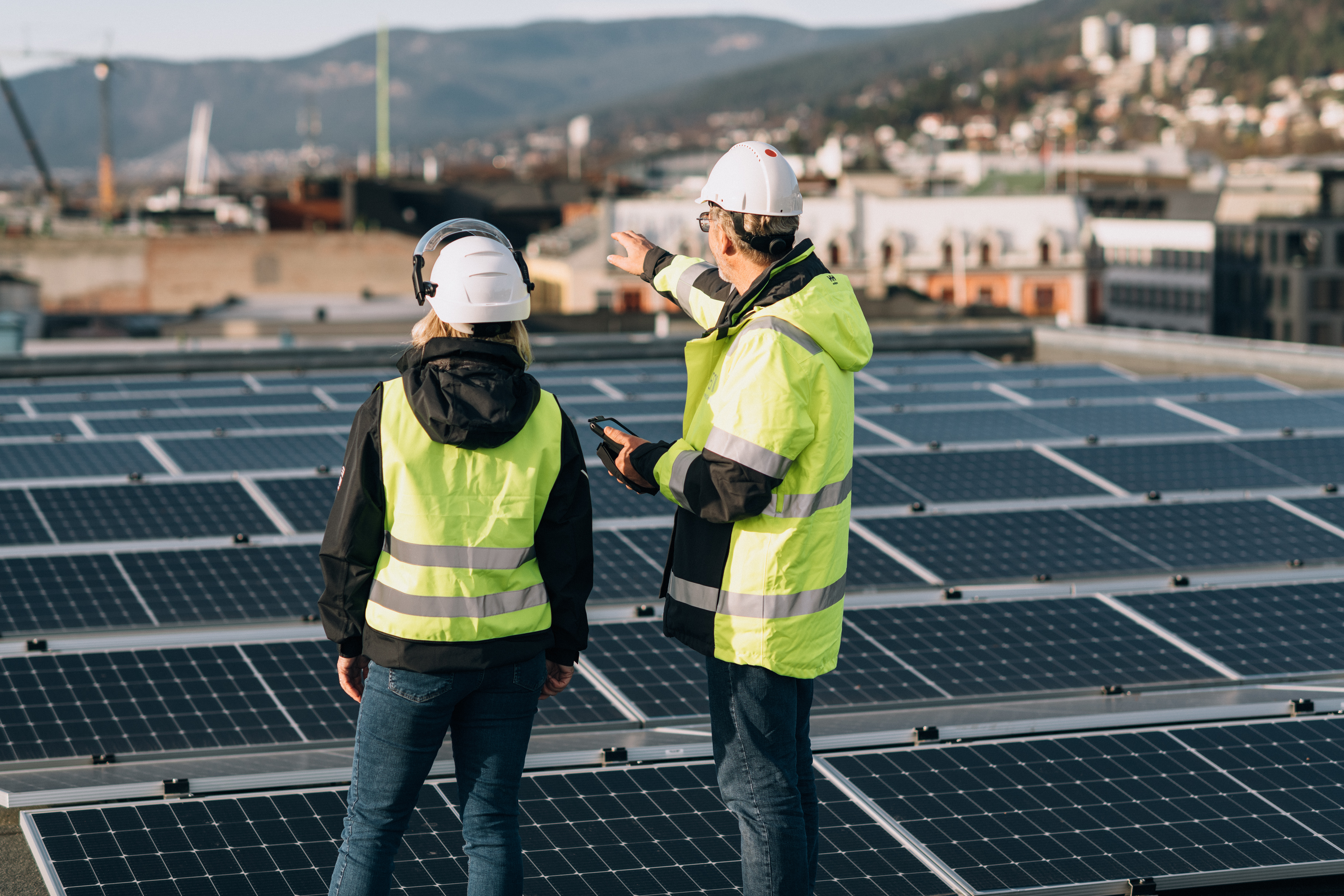 A man and a woman in safety gear standing on a rooftop with solar panels, pointing toward the cityscape with mountains in the background.