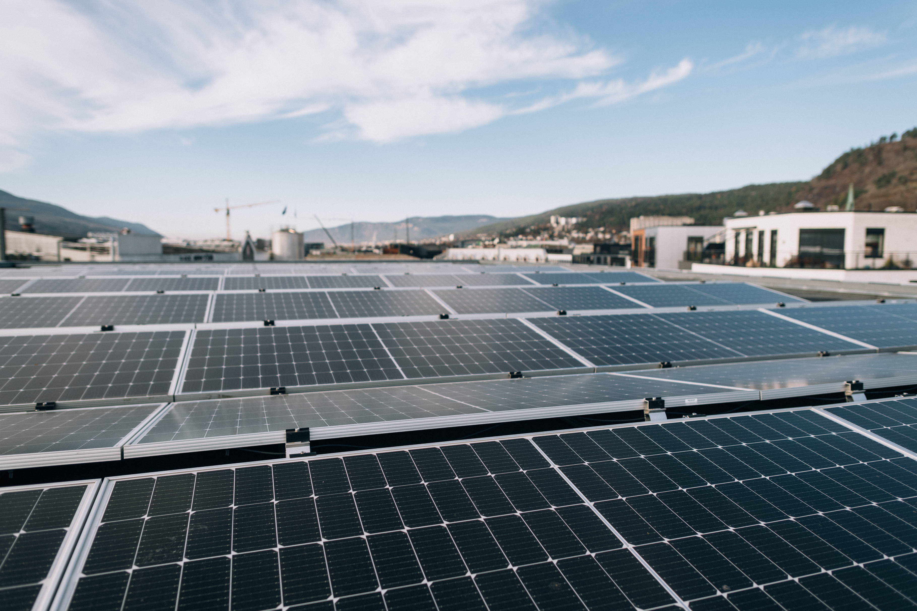 Rows of solar panels on a rooftop, with buildings, cranes, and hills in the background.