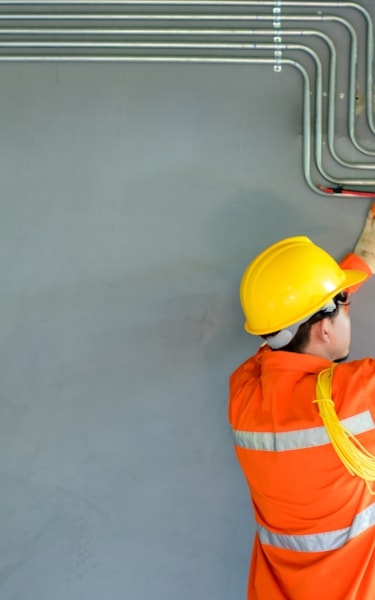 Person in safety gear working on an electrical panel with cables and conduits on a gray wall.