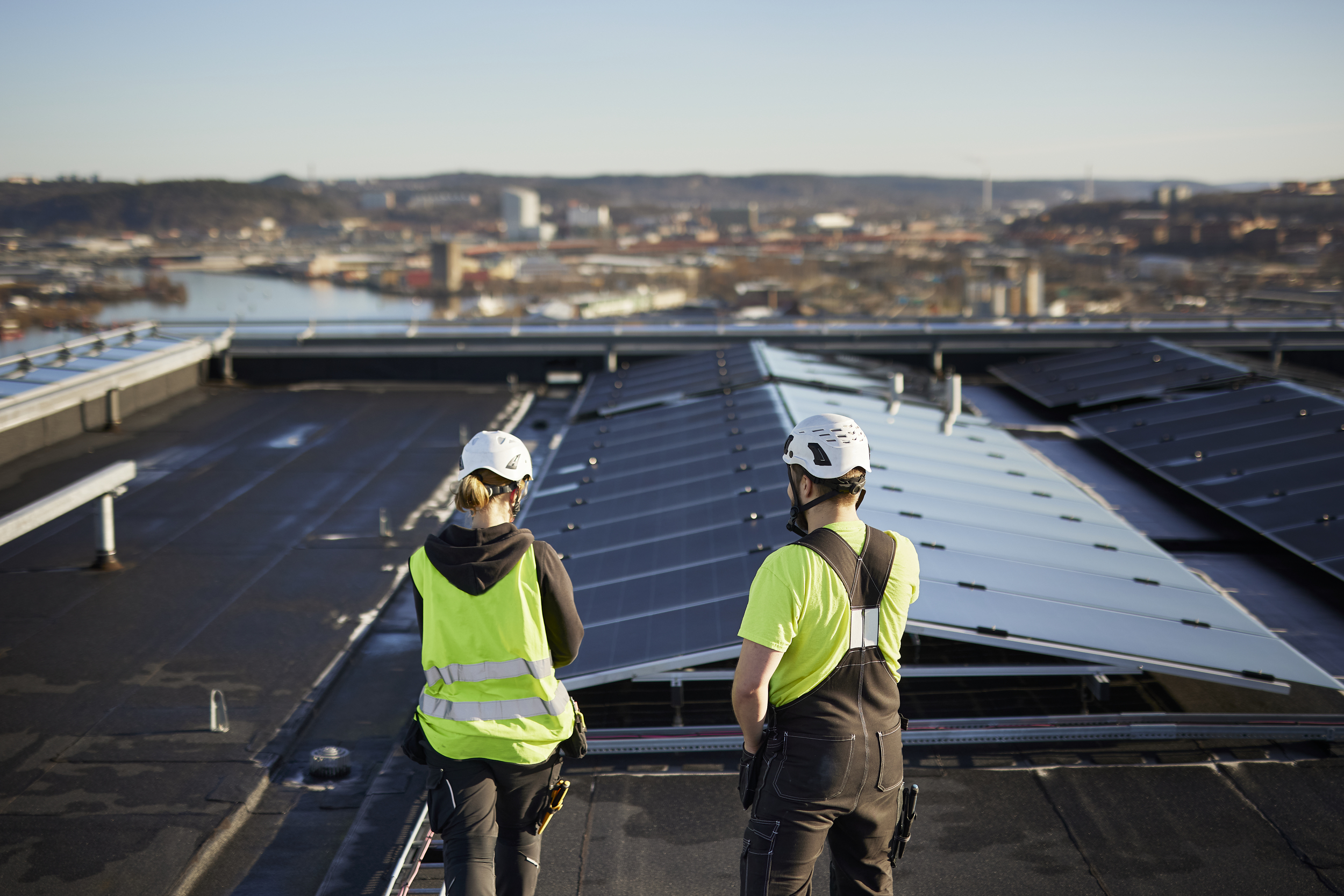 Two people in safety gear on a rooftop with solar panels, with a city and body of water in the background.