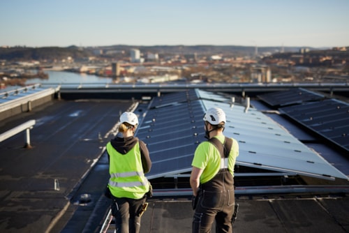 Two people in safety gear on a rooftop with solar panels, with a city and body of water in the background.